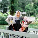 Smiling Muslim woman in hijab celebrates graduation outdoors with balloon and flowers.