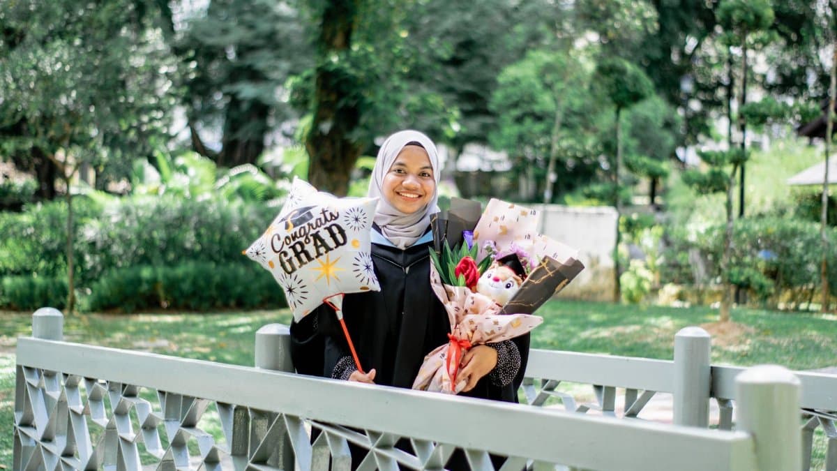 Smiling Muslim woman in hijab celebrates graduation outdoors with balloon and flowers.