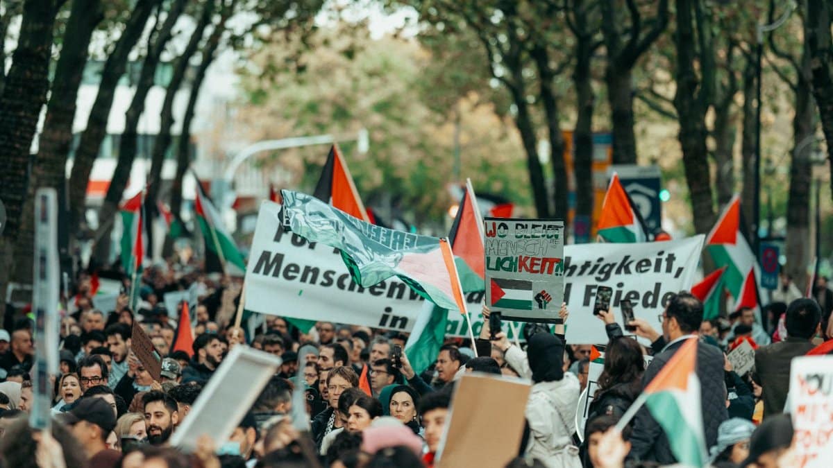A large, diverse crowd participates in a peaceful outdoor protest, raising flags and signs.