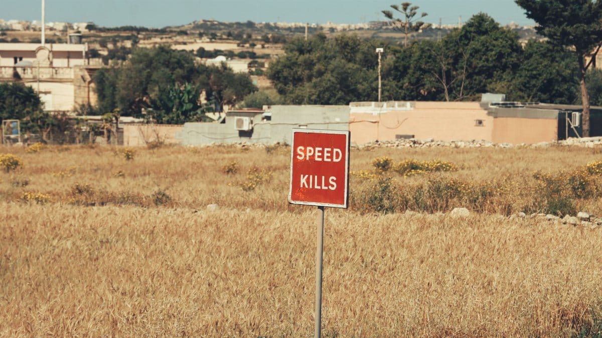 A red 'Speed Kills' sign stands in a sunlit Maltese field, highlighting road safety.