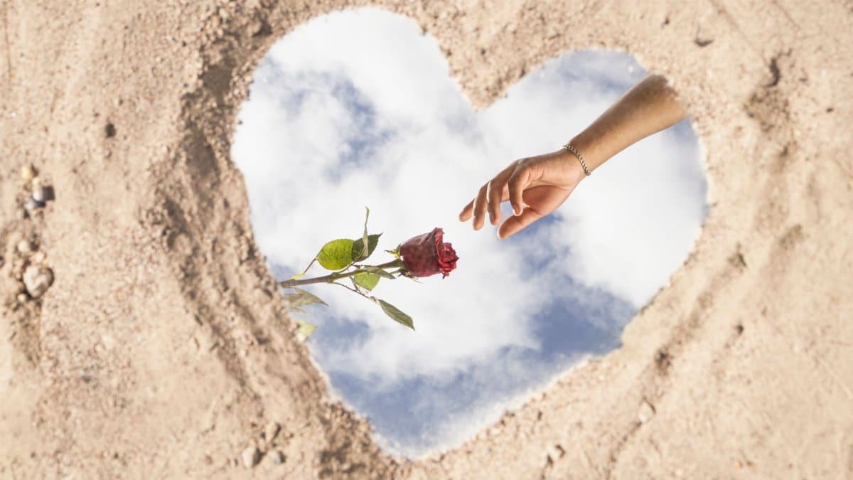A heart shape on sand reflects the sky, with a hand reaching for a rose, symbolizing love and connection.