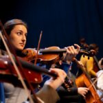 A group of musicians perform in an orchestra, featuring a violinist in focus.