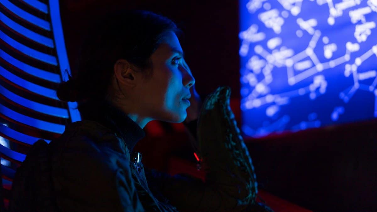 A woman in a space suit examines star maps in a futuristic, blue-lit environment, suggesting space exploration and technology.