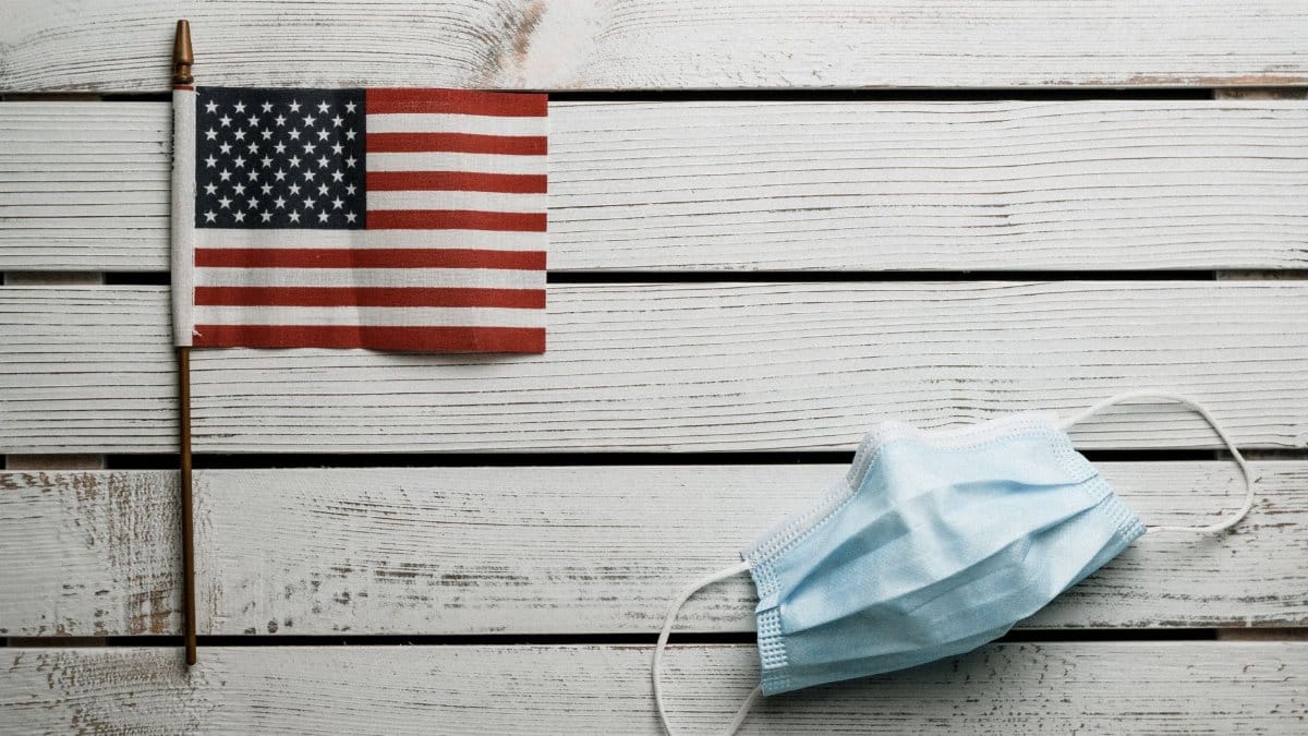 Top view of small Flag of United States and light blue fabric disposable face mask on white wooden wall