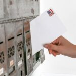 Close-up of a hand inserting a stamped envelope into a mailbox, symbolizing voting.