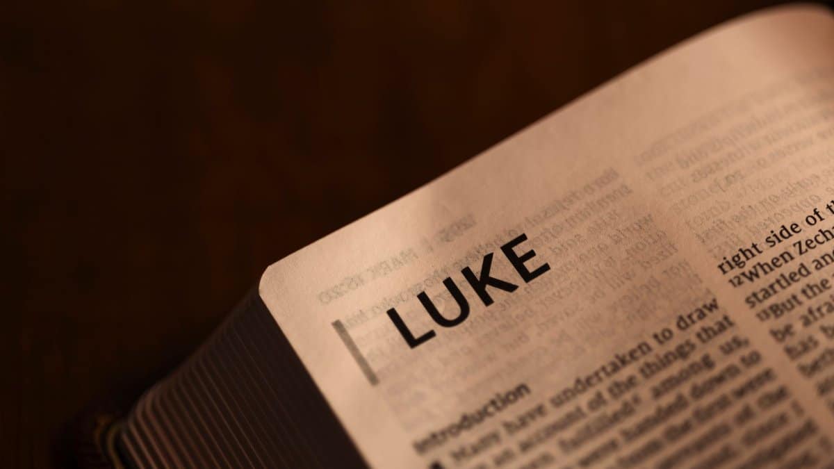Close-up of an open Bible showing the start of the Book of Luke, softly lit on a dark background.