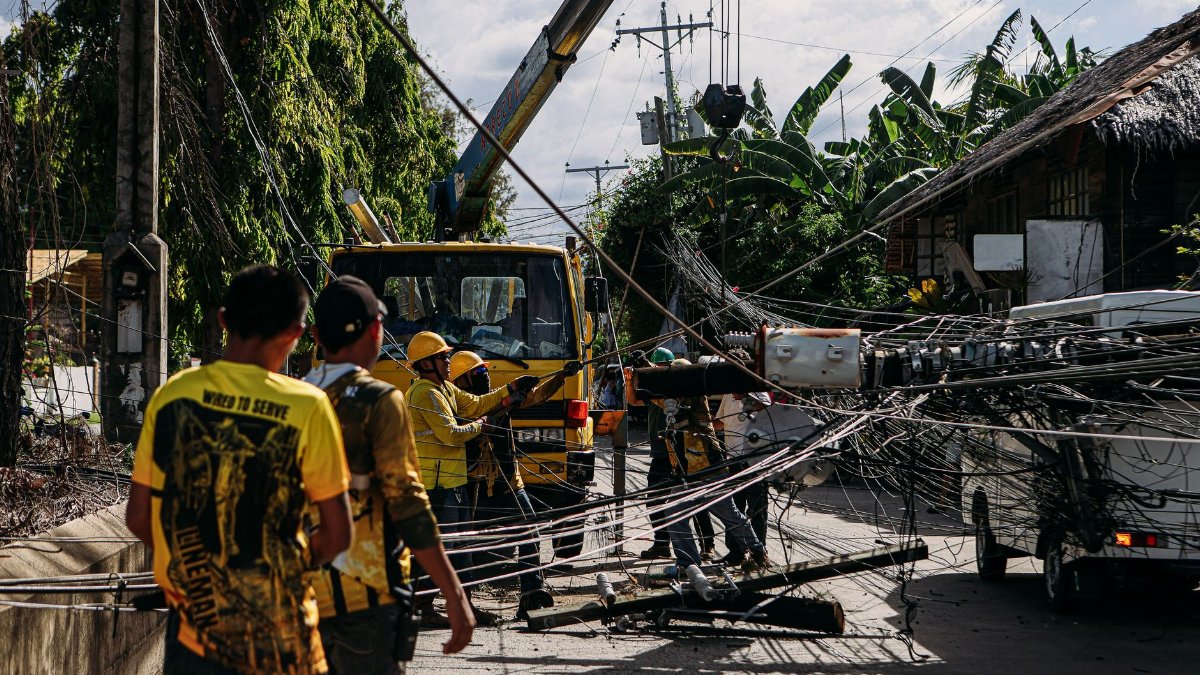 Workers fixing damaged electrical lines on a street post-storm, surrounded by fallen cables and equipment.