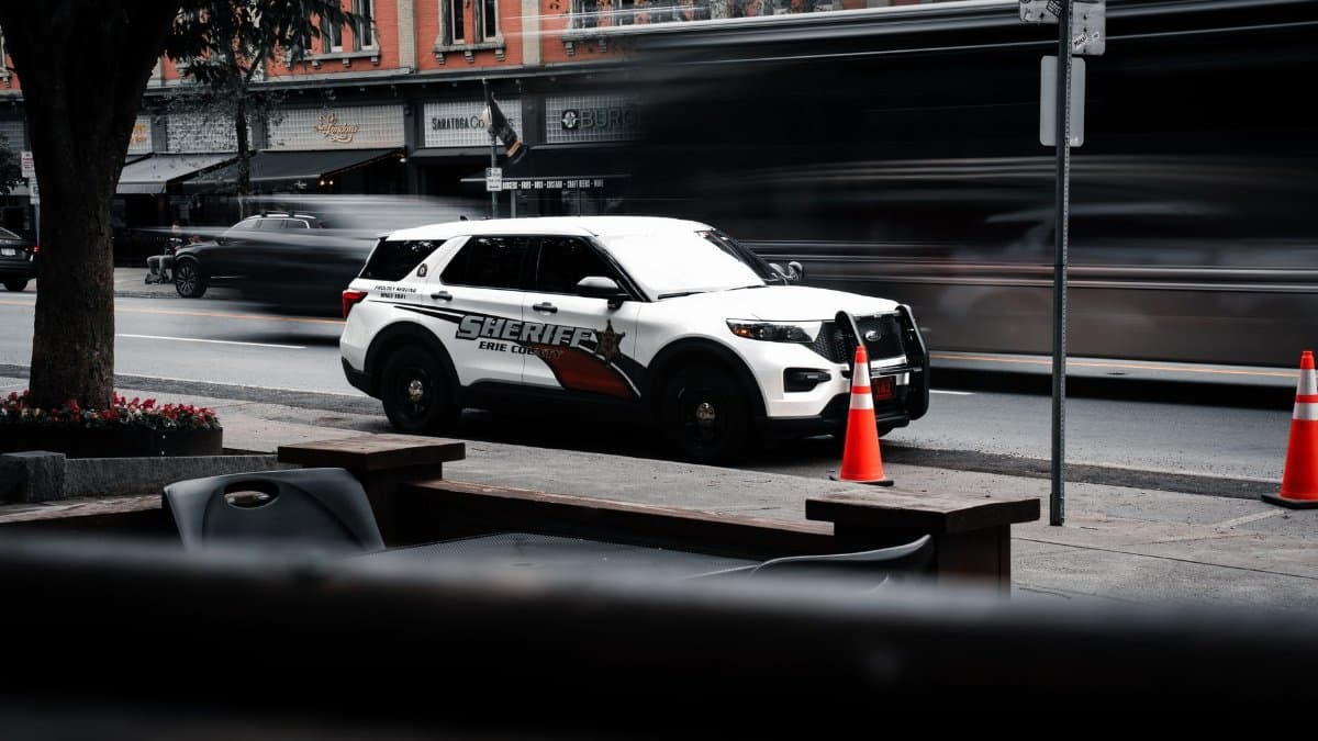 A sheriff vehicle parked on a busy urban street with motion blur of passing cars and vibrant orange cones.