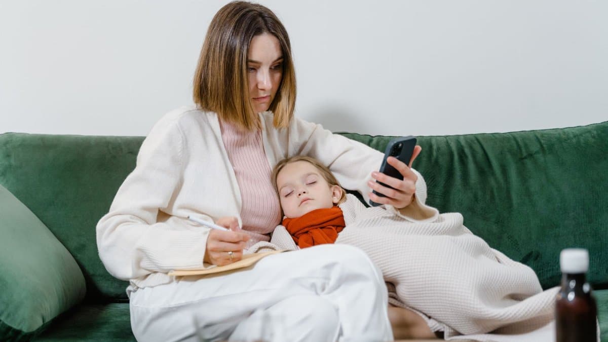 Woman researches symptoms on phone while comforting sleeping child on couch.