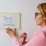 A teacher in pink blazer writing 'Back to School' on a small whiteboard indoors.