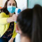Female teacher checks student's temperature with thermometer in classroom.