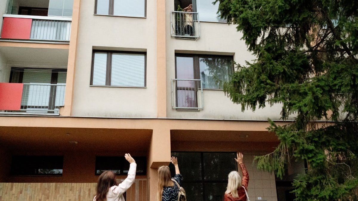 Three women waving to an elderly woman on an apartment balcony in Prague.