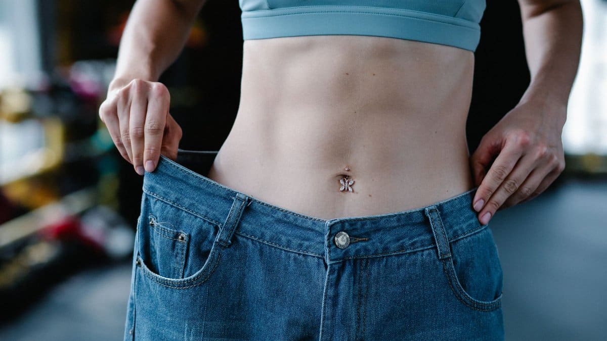 A woman shows her weight loss by holding oversized jeans revealing her toned stomach.