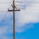 Wooden power pole with transmission lines against a clear blue sky and clouds.