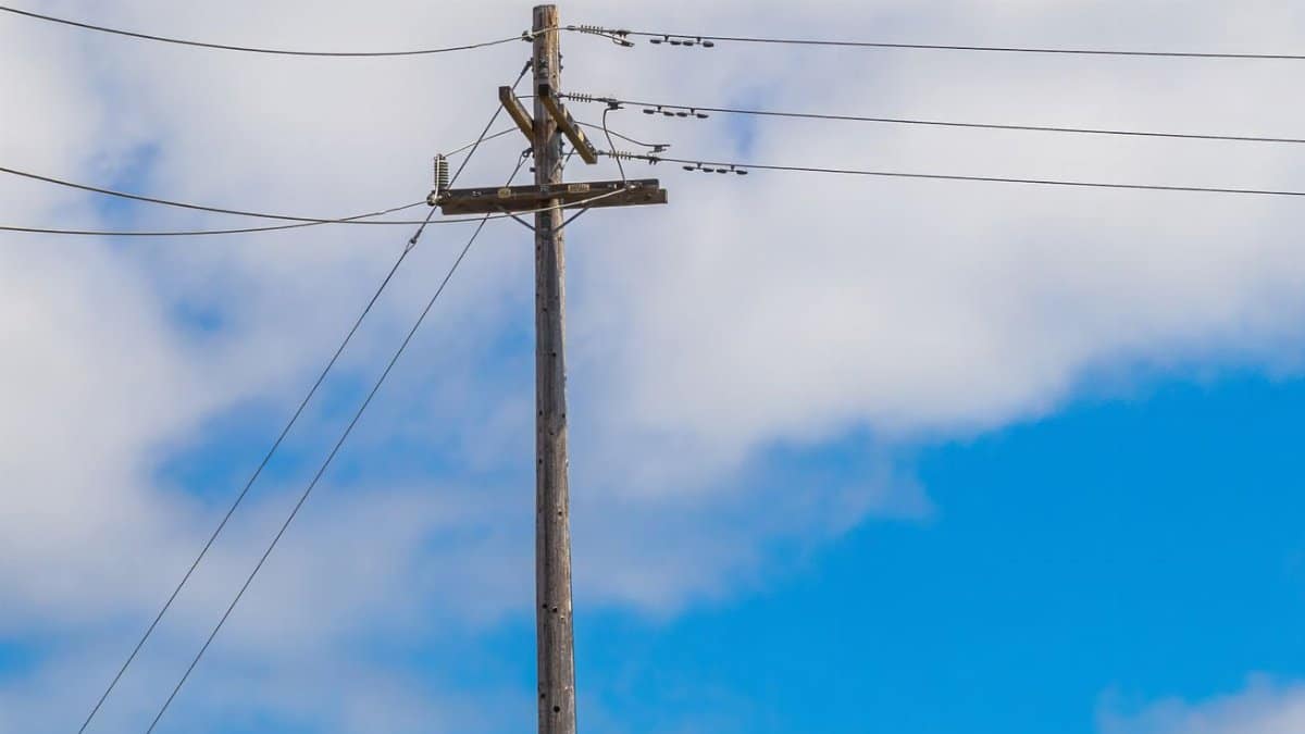 Wooden power pole with transmission lines against a clear blue sky and clouds.