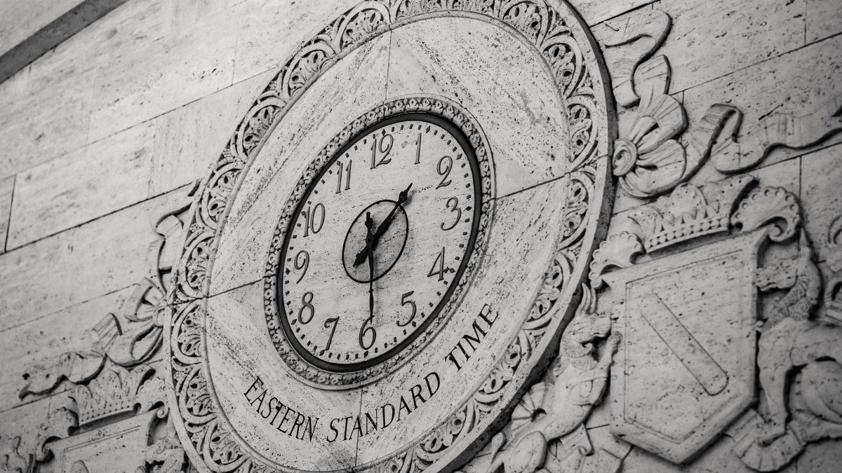 From below black and white of stone wall with ornamental details around clock showing Eastern Standard Time