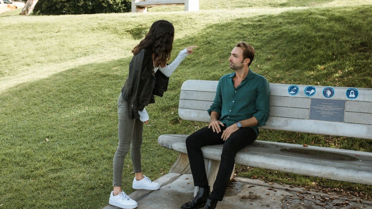 A couple in a heated argument while sitting and standing in a sunny park setting.