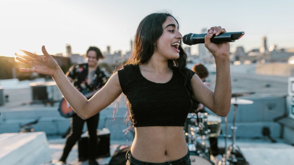 Young woman singing during a vibrant rooftop music session with band at sunset.