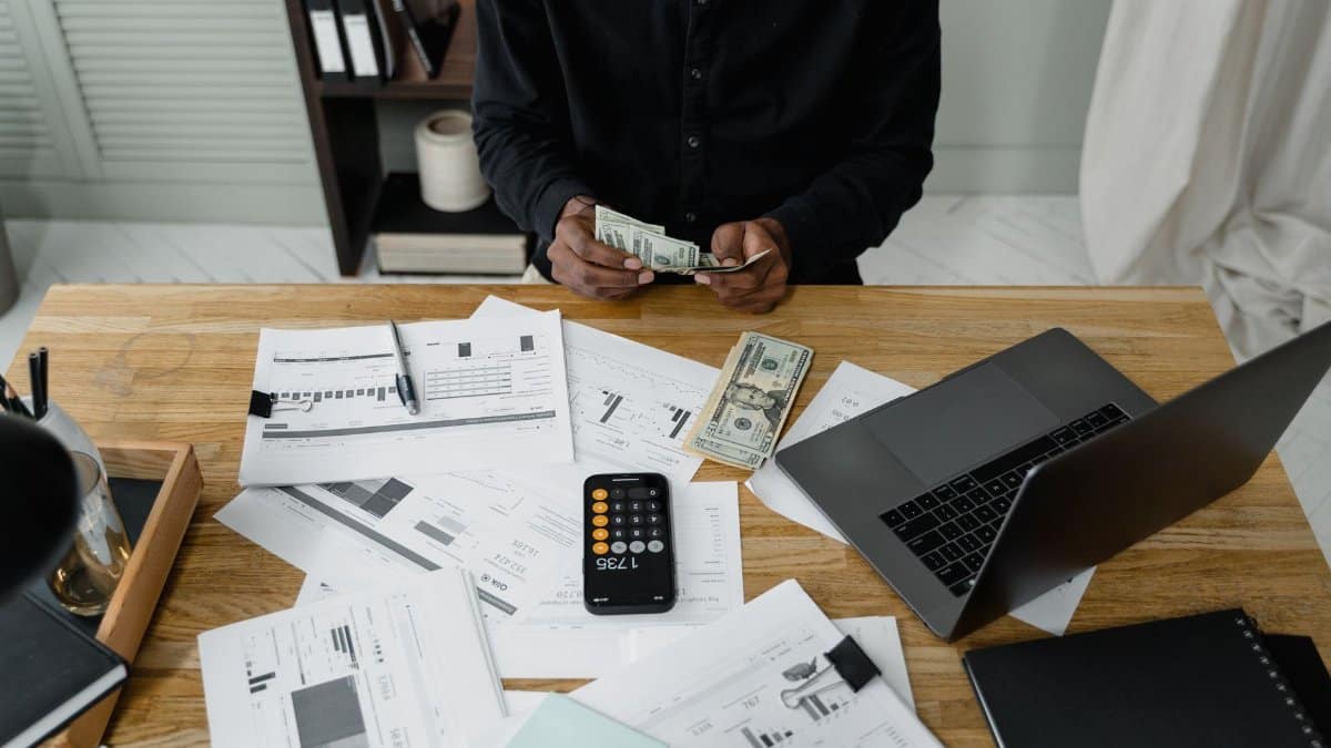 A person counts dollar bills at a desk with a laptop, papers, and calculator.