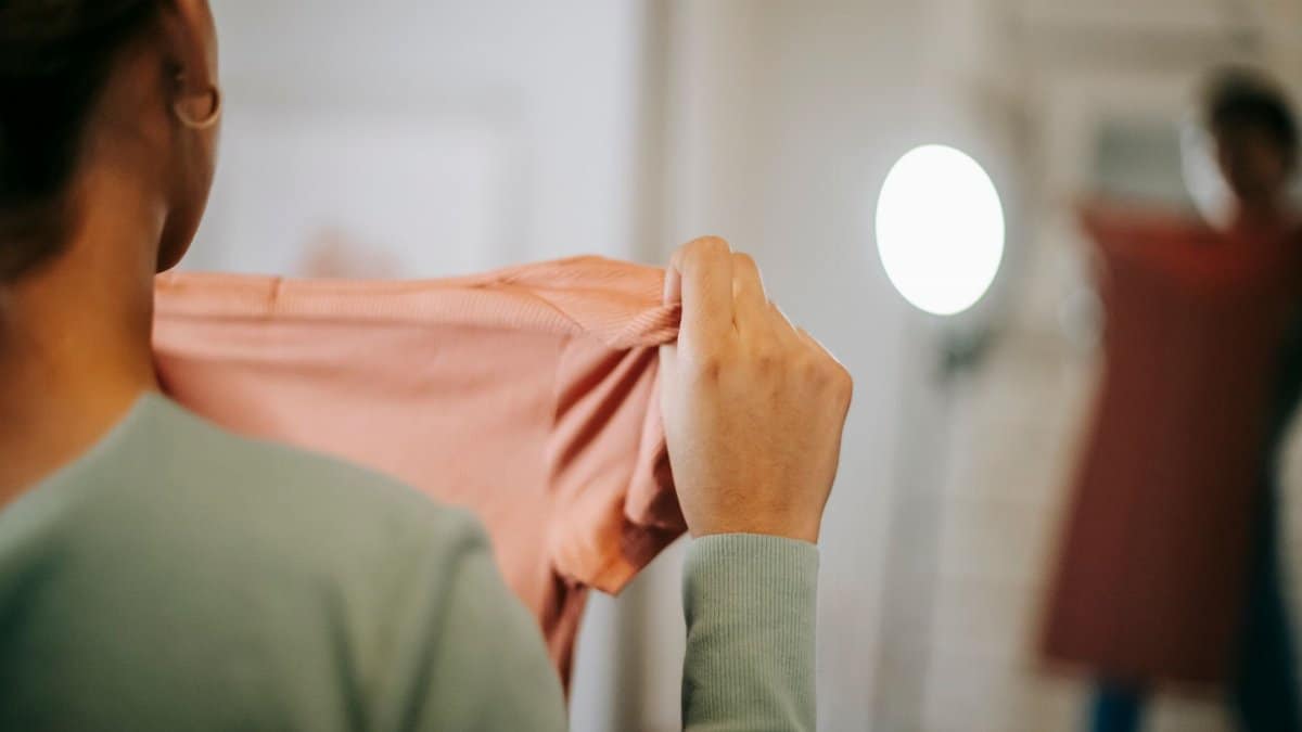 Back view of unrecognizable female in casual clothes showing pink dress while selecting clothes and looking in mirror while standing in light room at home
