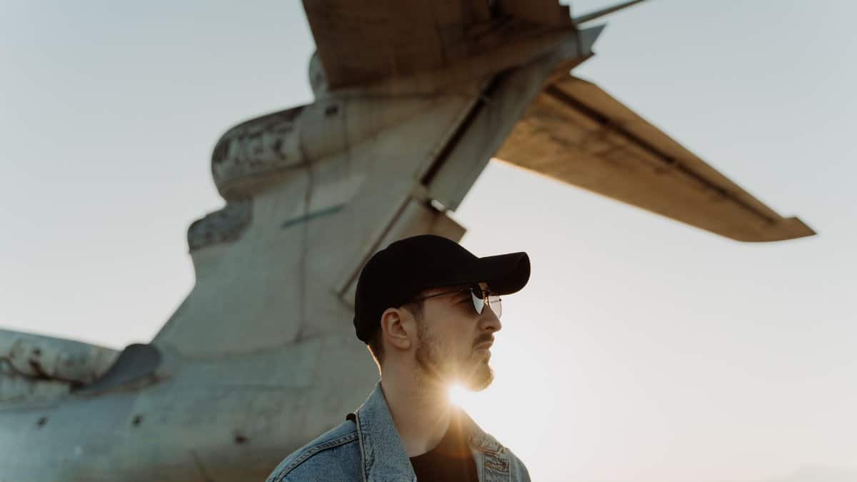 Man in denim and cap by aircraft tail at sunset, fashionably cool.