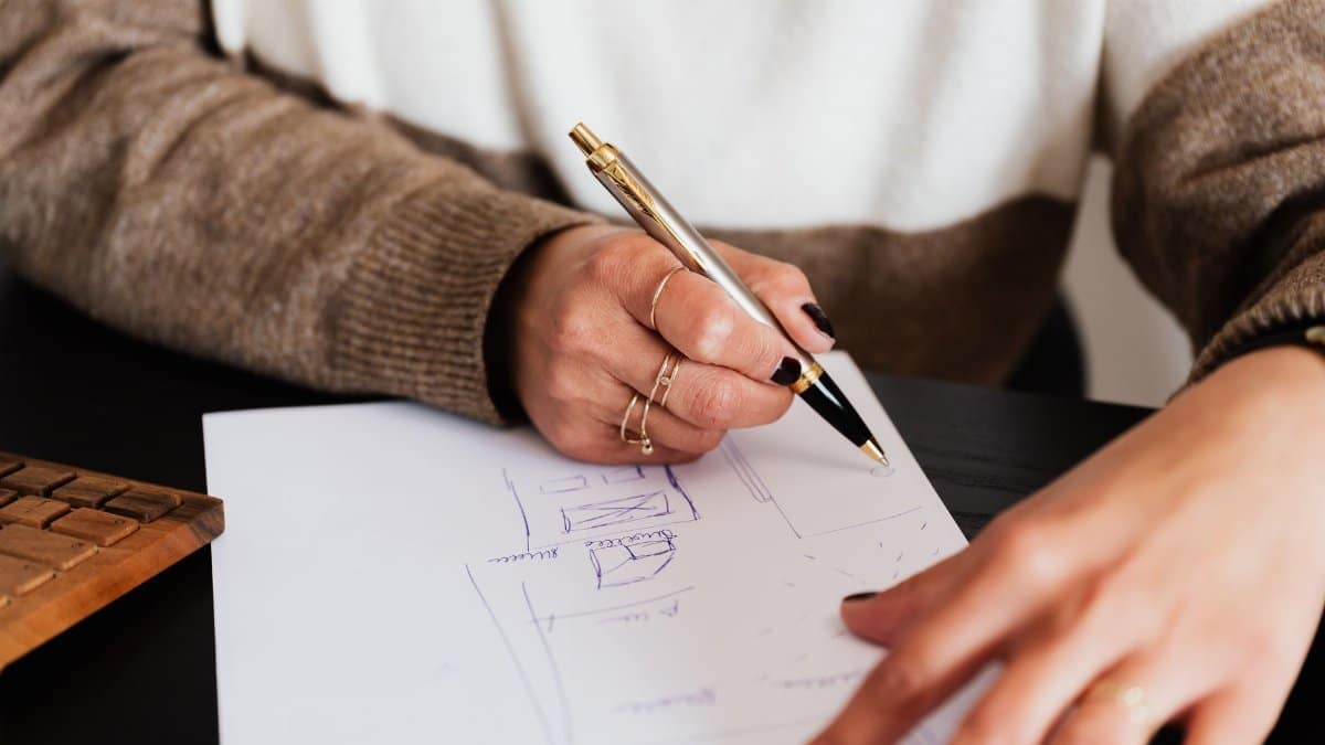 Crop anonymous elegant lady drawing draft plan of room with pen sitting at computer desk