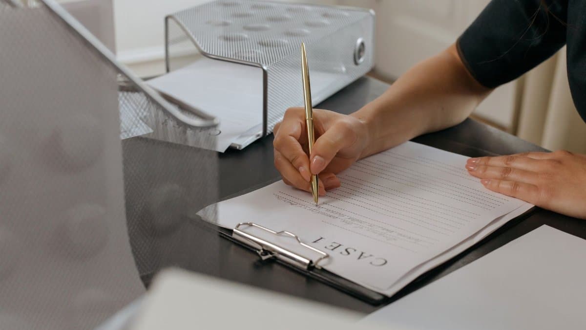 Close-up of a woman writing on documents with a gold pen in an office environment.