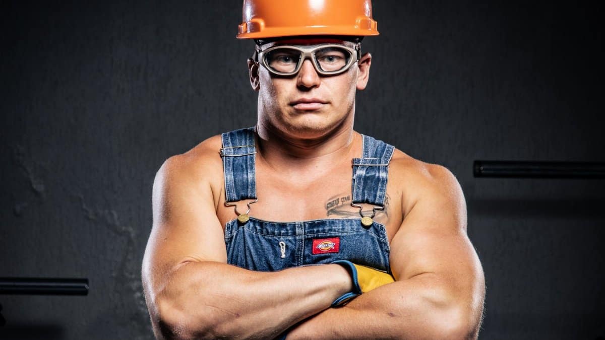 Portrait of a muscular construction worker in an orange hard hat and goggles with arms crossed.