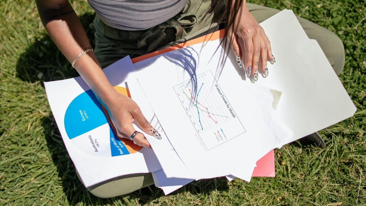 A woman sits on grass reviewing charts and business documents in a park setting, capturing an outdoor work scene.