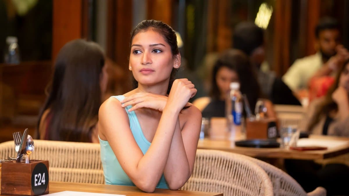 A young woman in a turquoise top sits in a lively, crowded restaurant, appearing thoughtful.