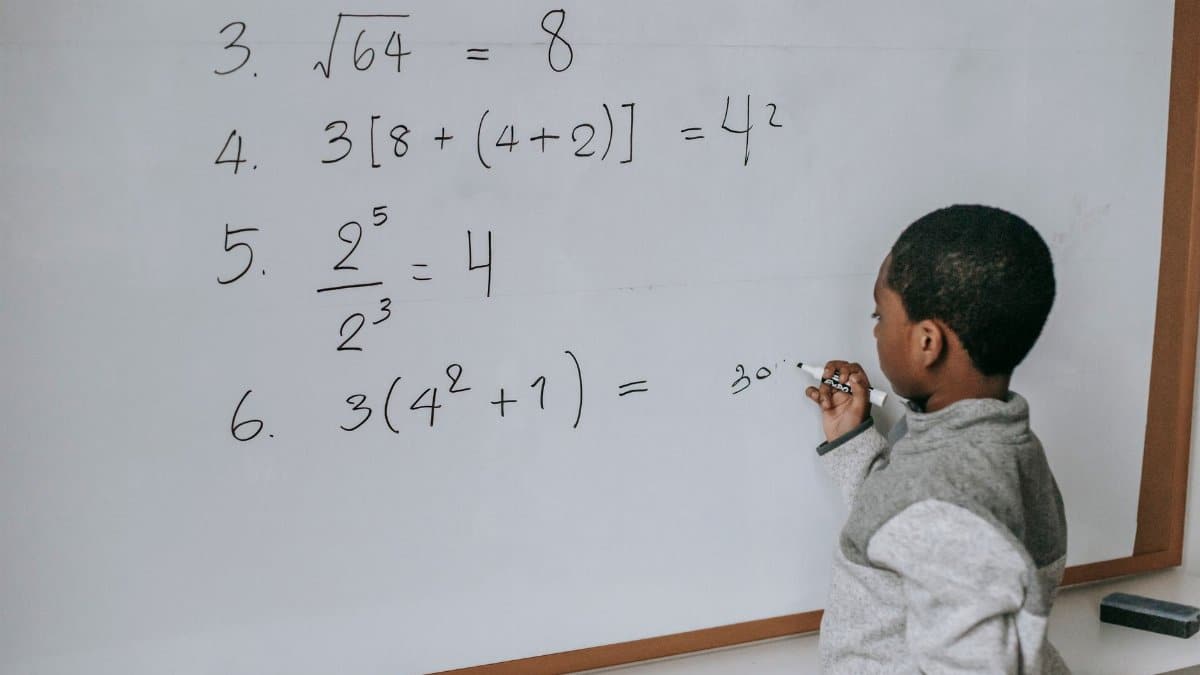 Side view of concentrated African American pupil standing near whiteboard in classroom and solving math examples during lesson at school