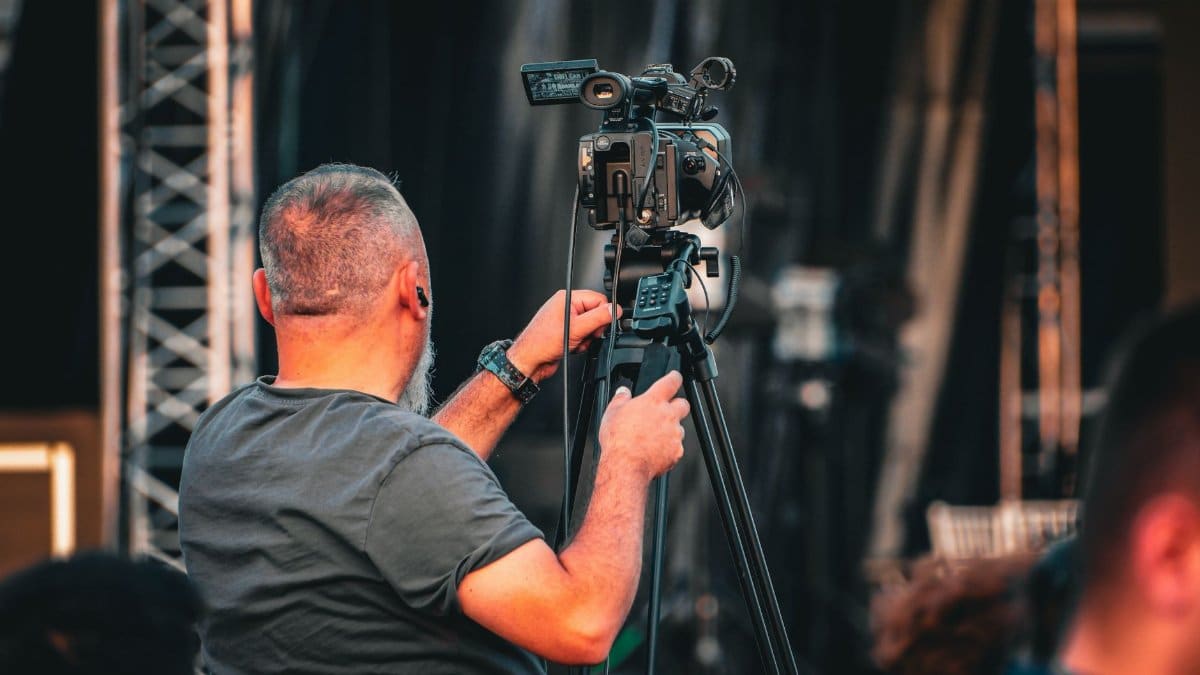 A professional cameraman operating a video camera on a tripod during an outdoor event, captured from behind.