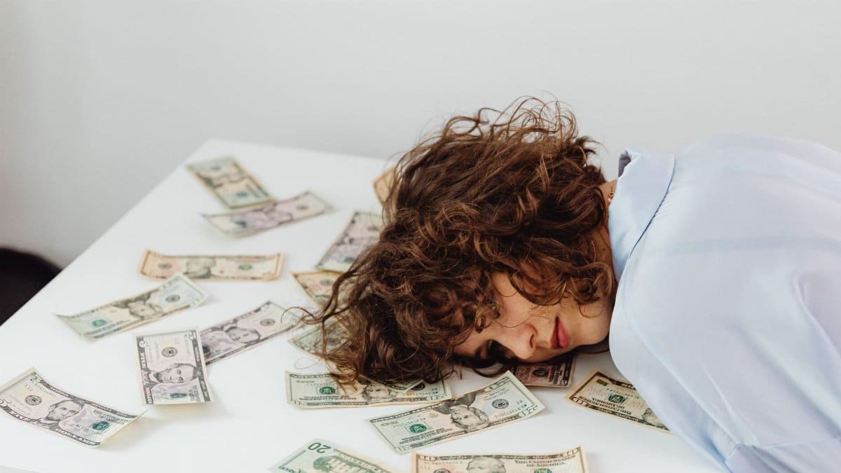 A woman with curly hair resting her head on a table covered in US dollar bills.