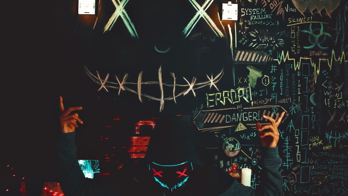 A masked individual stands before a chalkboard filled with neon drawings and cryptic messages, creating a mysterious ambiance.
