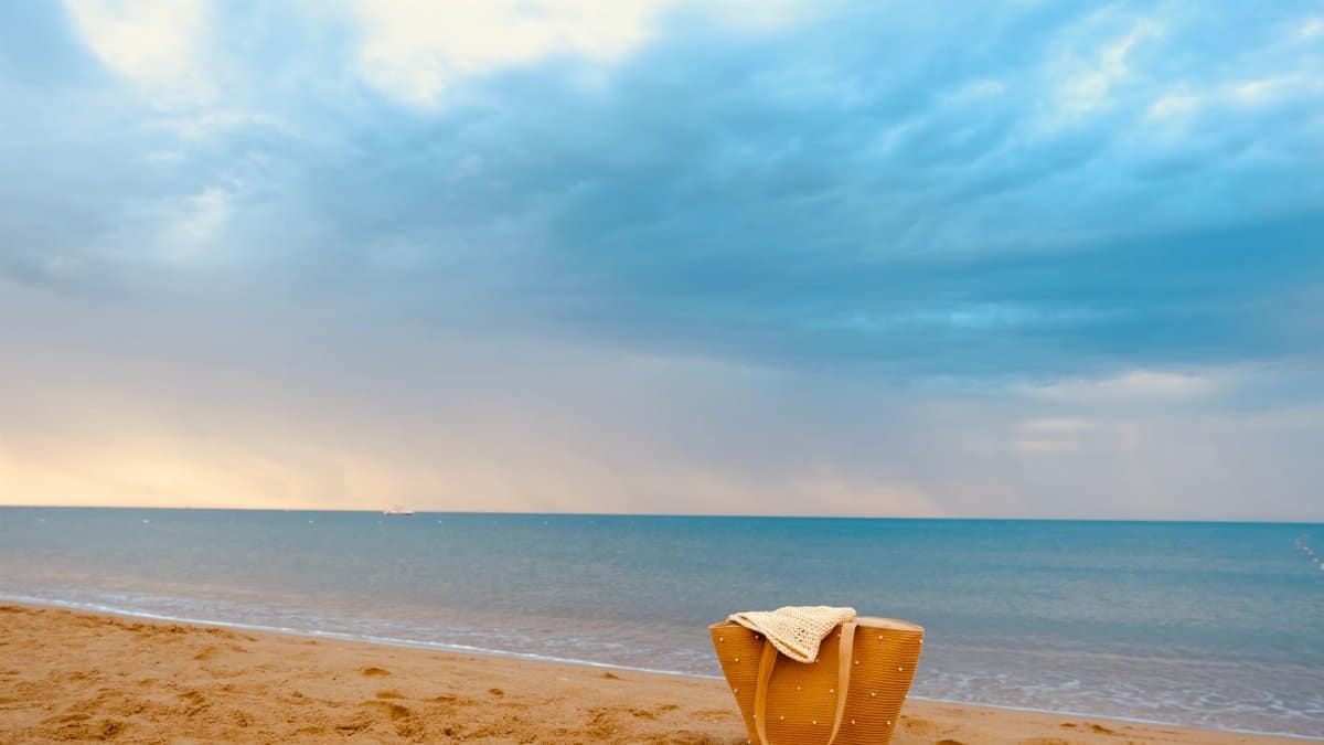 Peaceful beach scene with a straw bag on the sand against a cloudy sky and calm sea.