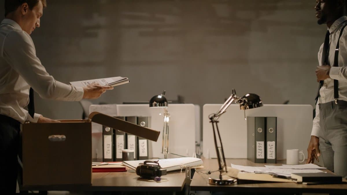 Two office workers sorting documents in a dimly lit workspace with several file binders.