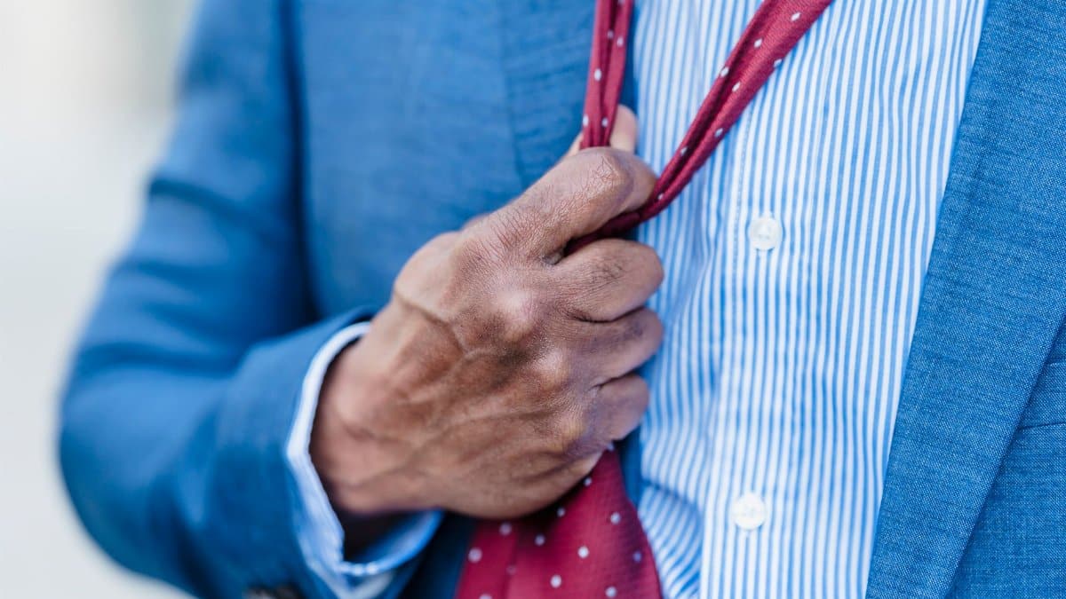 Crop anonymous tired male entrepreneur in formal suit adjusting red tie standing on street in city against blurred background after workday