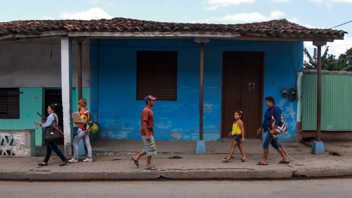 A diverse group of people walking on a sidewalk in front of colorful houses with a clear blue sky.