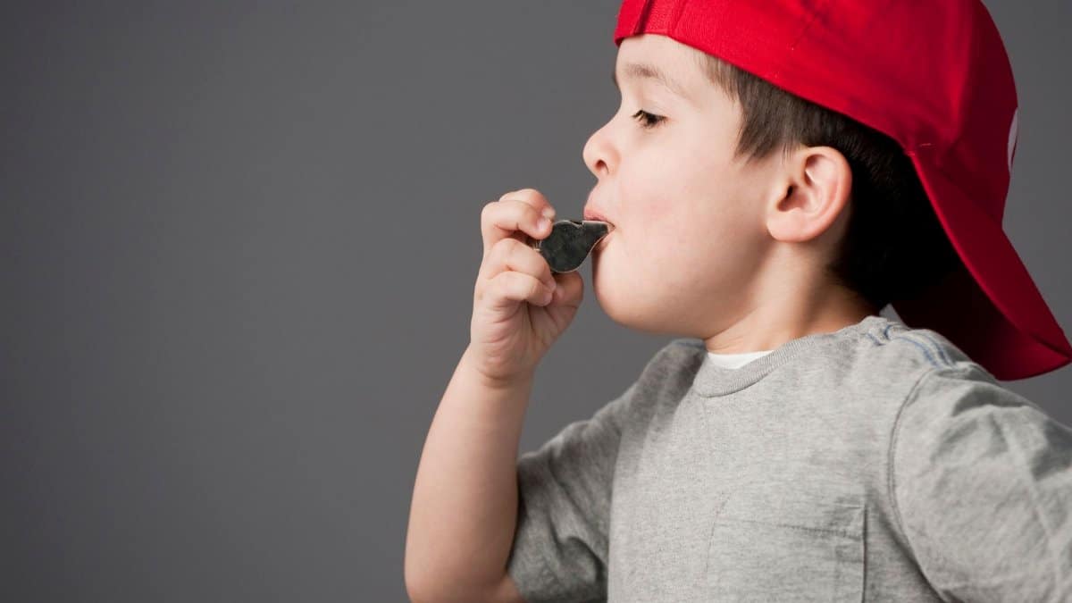 Side profile of a child with a red cap blowing a whistle against a gray background.