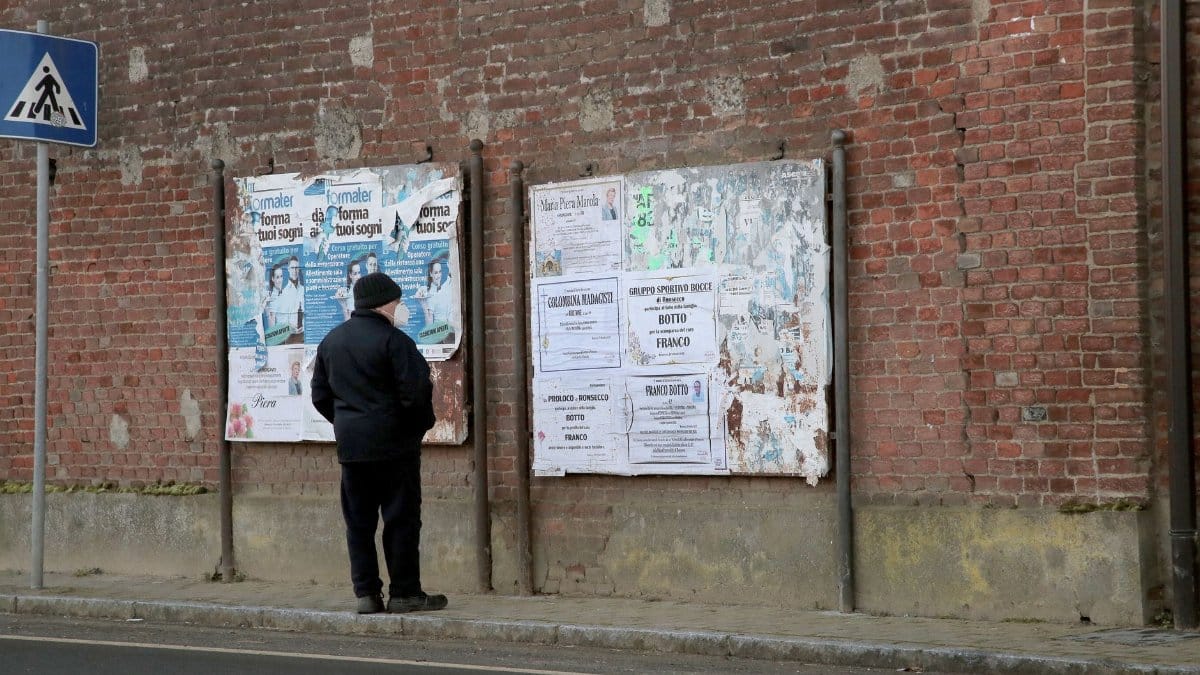 A man reads public notices on a brick wall along an urban sidewalk.