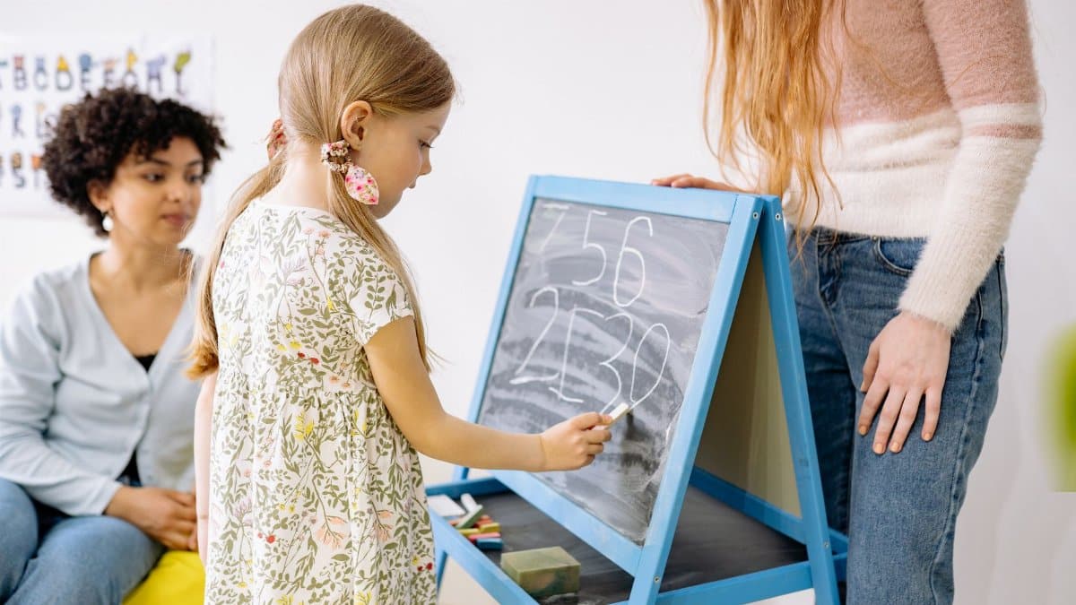 A young girl writes numbers on a chalkboard in a classroom, supervised by adults.