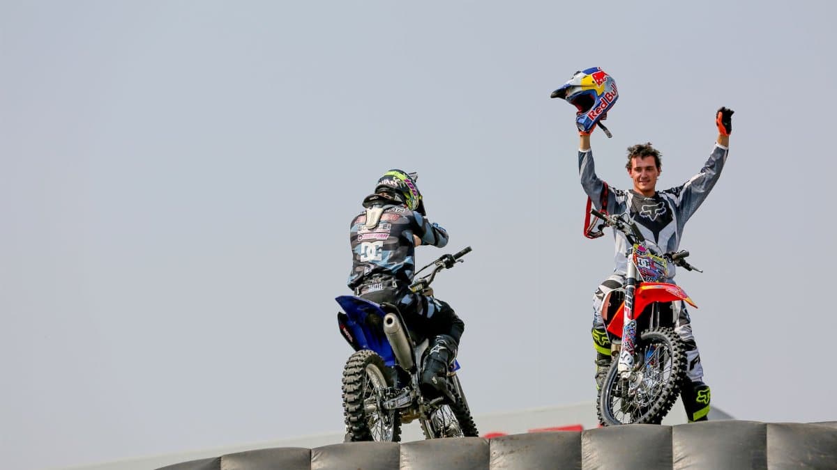 Two motocross riders on bikes, one raising a helmet victoriously outdoors.
