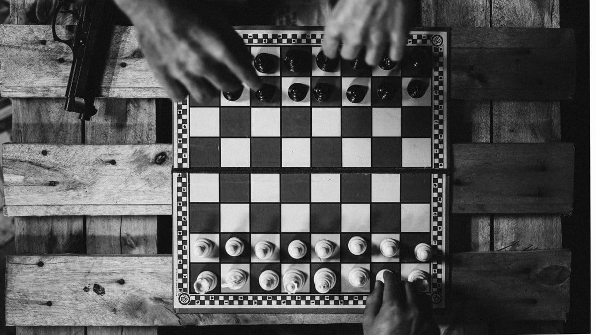Black and white chessboard setup with hands moving pieces on a wooden table alongside a gun.
