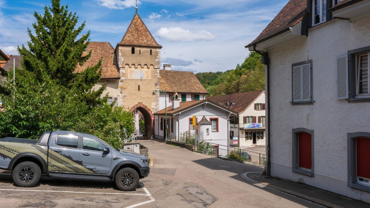 A picturesque view of a medieval gateway and a parked truck in a European town, under a clear sky.