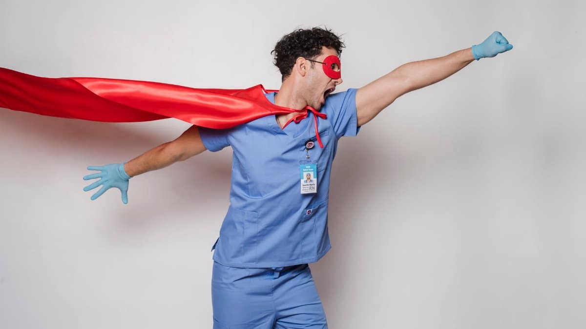 Emotional male medic with open mouth in uniform and fluttering red cape with mask standing on white background with outstretched arms