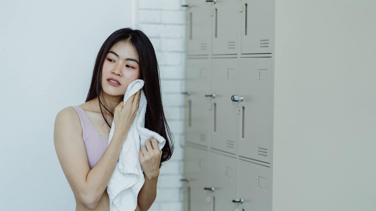 Woman in a locker room drying off with a white towel, next to lockers.