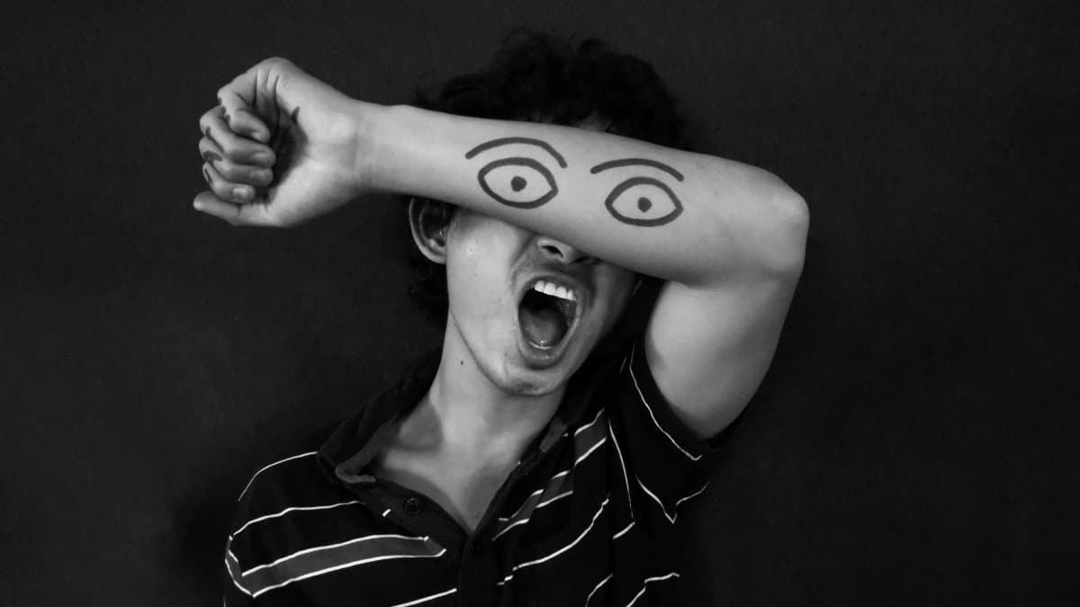 A person covers their eyes with an arm featuring drawn eyes, expressing creativity and emotion.