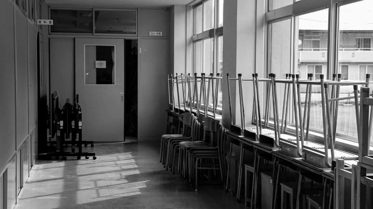 Black and white photo of an empty school corridor with stacked chairs and sunlight streaming through windows.