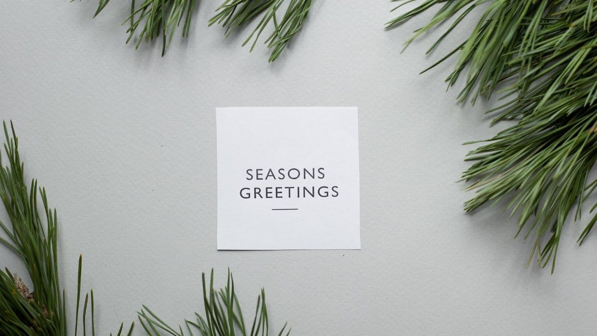 Top view of Season Greetings phrase written on greeting card placed on gray background with green spruce branches in studio