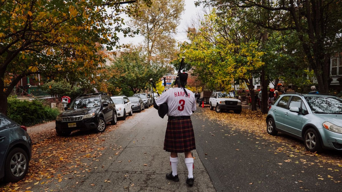 A man in a kilt plays bagpipes while walking along a leaf-covered city street in autumn.